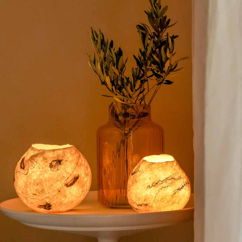 Les cocons lumineux en laine feutrés de la Ferme du Lambert sur table blanche avec vase et branche d'olivier.